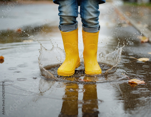 A child's feet in yellow rain boots splashing in a water puddle.