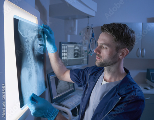 A medical professional in blue gloves examining an orthopedic X-ray of the cervical spine in a radiology room, with digital monitors and medical equipment in the background.
