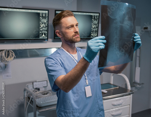 A medical professional in blue gloves examining an orthopedic X-ray of the cervical spine in a radiology room, with digital monitors and medical equipment in the background.