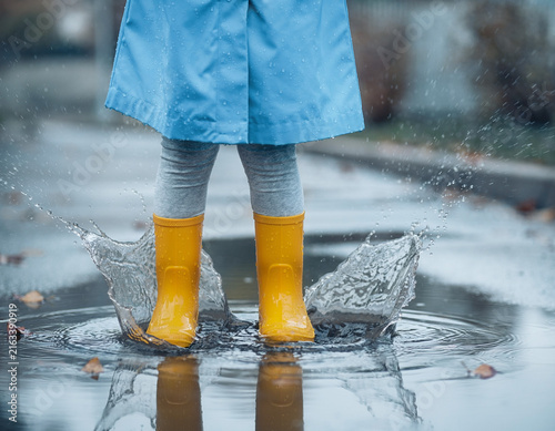 A child's feet in yellow rain boots splashing in a water puddle.