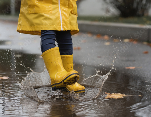 A child's feet in yellow rain boots splashing in a water puddle.