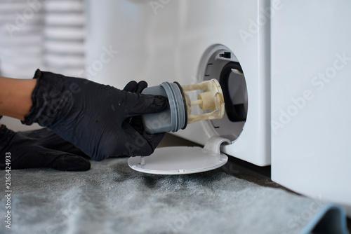 Konstfotografi Gloved hand removes drain pump filter from washing machine through lower access