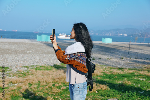 Foto Woman taking photo on a sunny beach with phone held up, windswept hair, casual jacket, jeans, coastal landscape, distant boats, calm sea, vibrant sky, candid travel moment