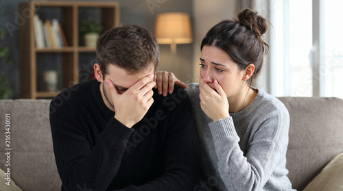 Distressed young couple comforting each other while sitting on sofa at home, representing grief, emotional crisis, relationship support, sadness and coping together.