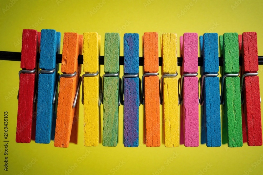 Row of colorful wooden clothespins clipped onto black cord against bright yellow background. Simple items arranged in neat line, with unique vibrant hue. Practical household objects used for hanging