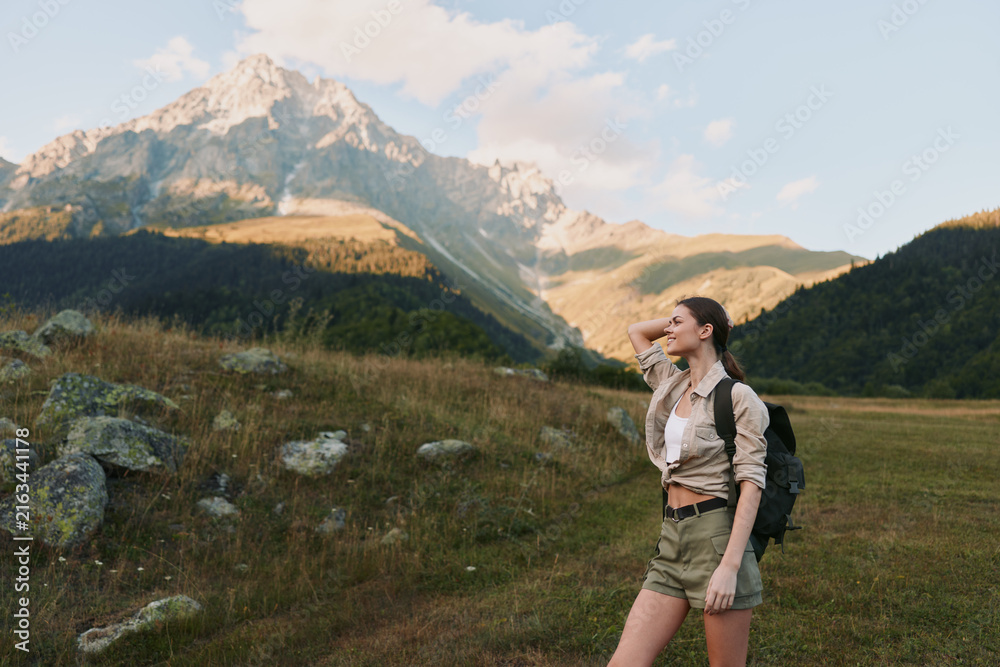 Naklejka premium Young woman hiker with a backpack stands on a meadow, looking toward distant mountains under clear sky, capturing an outdoors lifestyle moment of nature, adventure, and travel
