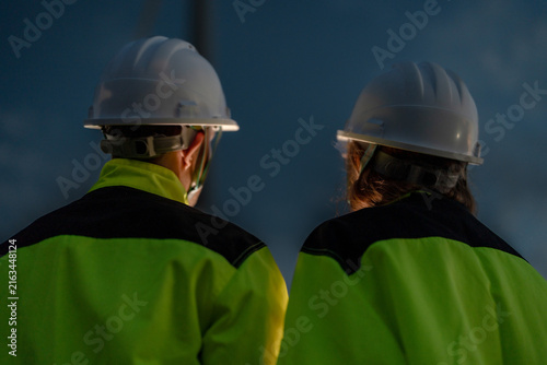 Bild auf Leinwand Two wind energy professionals seen from behind stand quietly as blades sweep the darkening sky on high ground overlooking the wind farm in burnt orange afterglow at a wind power plant in