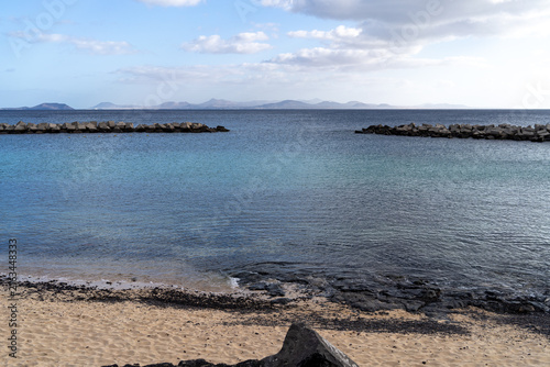View of the serene Lanzarote coastline, featuring turquoise water, sandy beach, and breakwaters under a cloudy sky.
