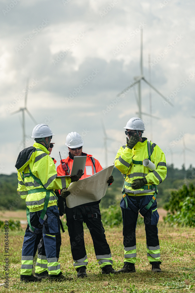 Fototapeta premium A mixed team of wind farm engineers in yellow and orange hi vis debate cable routing over a large site plan on a green meadow with rows of turbines behind in bright overcast daylight on a.