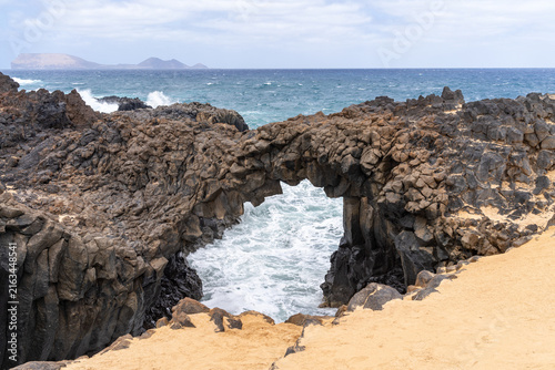 A natural rock arch with waves crashing underneath, framed by a sandy beach. La Graciosa island in the distance.