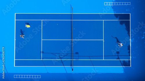 an empty blue hard tennis court scattered with yellow balls after a training session. The net, crisp white lines, and long shadows create a striking geometric background.