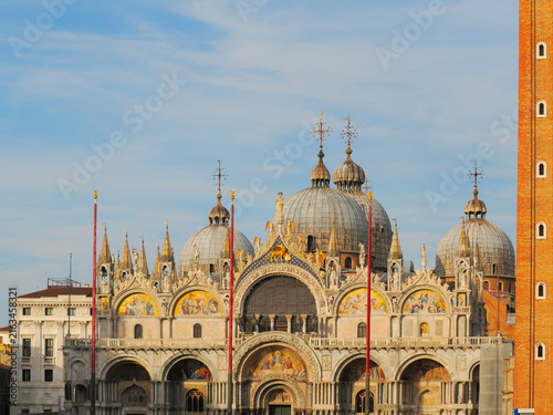 Ornate facade of St Mark's Basilica with golden domes and bell tower against blue sky in Venice