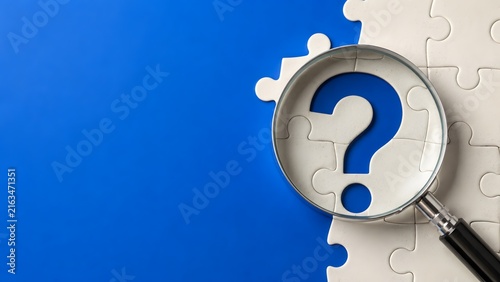 Magnifying glass over a white jigsaw puzzle with a question mark on a blue background representing problem solving