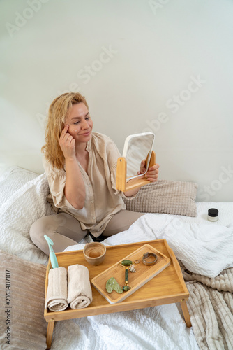 Woman in early forties holding mirror looking wrinkles on her face, morning beauty routine 