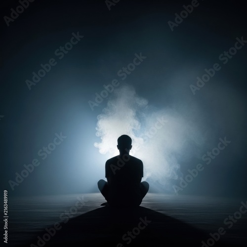 Silhouette of a person sitting in meditation pose against bright backlighting in a dark atmospheric room filled with dense fog and smoke, cinematic wide angle composition.