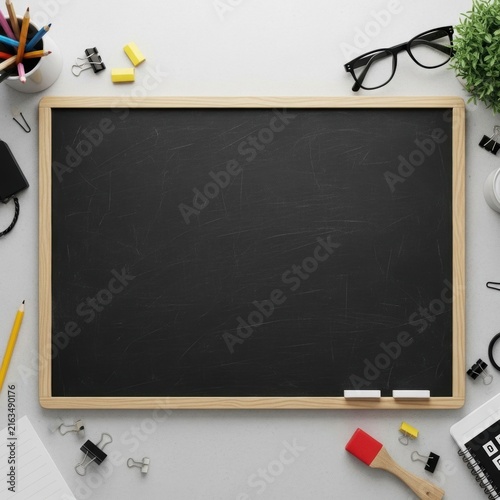 Top down flat lay view of a blank black chalkboard with a wooden frame, positioned on a light gray desk surrounded by school supplies, colored pencils, binder clips, and eyeglasses.
