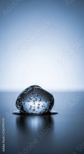 Vertical macro shot of a clear viscous liquid droplet containing small oxygen bubbles, sitting on a dark reflective wooden surface with a soft blue and white gradient backdrop.