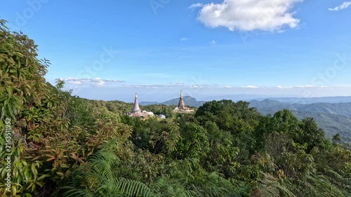 Wide shot of two beautiful Buddhist temples with sharp spires amidst dense green forest and mountains in northern Thailand on a sunny day.