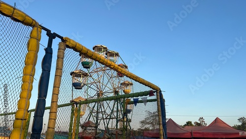 Fotografie Outdoor amusement area with a Ferris wheel and colorful attractions under a clear blue sky, viewed from a wide distance