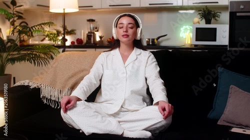 Young woman at night home wears headphones plays rain sound meditates deep breath slow calm finds peace. Caucasian girl on sofa at evening eyes closed quiet mood relax chill soft focus mindful rest.