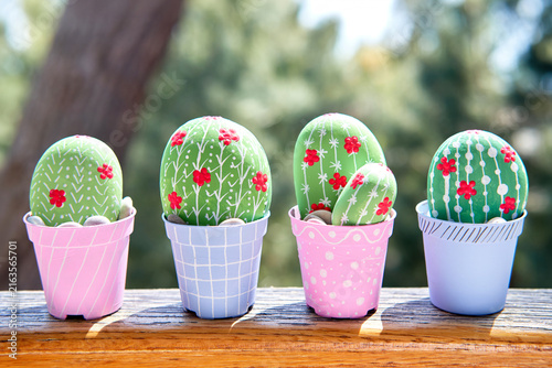 Four hand-painted stone cacti with vibrant red blooms and delicate white patterns sit in colorful pots on a sunlit wooden table; the soft outdoor backdrop and warm lighting highlight this charming DIY