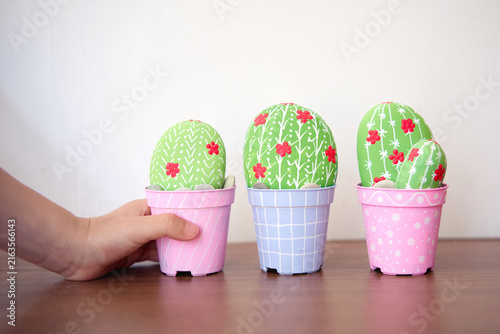 A child’s hand gently holds a miniature painted cactus in a pink striped pot; beside it, two more stylized cacti sit in pastel pots, adorned with red flowers, creating a cheerful DIY craft display.