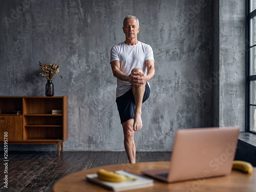 Focused senior man practicing a balancing yoga pose at home, enhancing flexibility, strength, and mindful wellness.