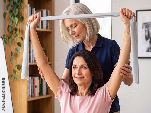 Professional physical therapist assisting a smiling mature woman with resistance band exercises for arm rehabilitation.