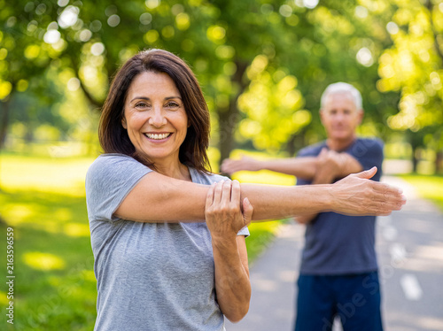 Foto Mature woman smiling and stretching arm in green park with man in background, engaging in outdoor fitness activity