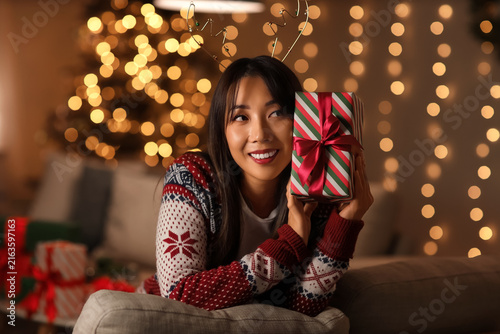 Beautiful young happy Asian woman with Christmas gift box sitting on sofa in ...