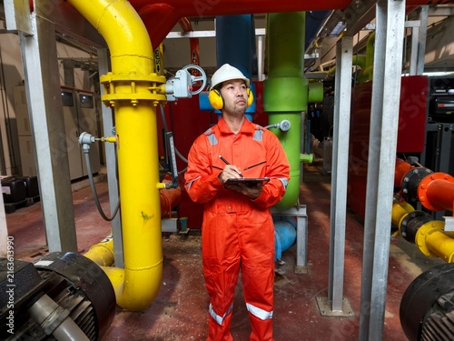 Obraz na płótnie An industrial engineer in an orange jumpsuit, safety helmet, and ear protection looking up while recording data on a digital tablet, surrounded by gas and cooling pipeline systems