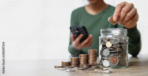 Woman putting coin into glass jar for saving money, with stacks of coins and smartphone on table. Financial planning and investment concept.