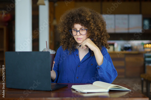A focused young woman with curly hair and glasses works on her laptop at a wooden cafe table, holding an iced beverage, ideal for remote work and freelance concepts