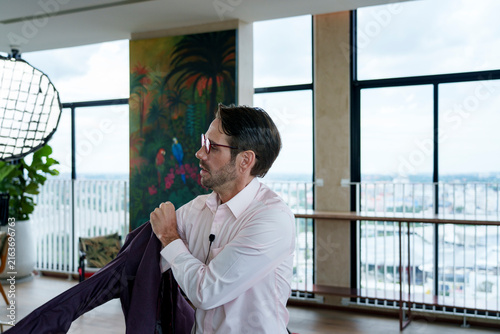 Obraz na plátně A white man with glasses putting a wine-colored jacket on in a well-lit room wit