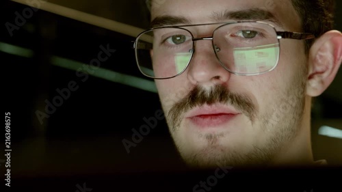 Close up of man wearing glasses with screen reflection visible in lenses. He appears concentrated, looking at computer monitor in dim environment