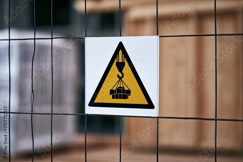 Cautionary sign depicting a crane lifting heavy loads, affixed to a metal fence