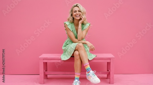 Happy young woman in a green dress and rainbow socks sitting on a pink bench against a bright pink studio background