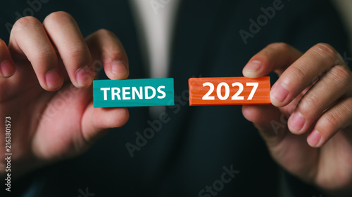 A professional in business attire holds blocks with the words Trends and 2027 in a modern office setting, symbolizing future planning, strategic thinking, and innovation.