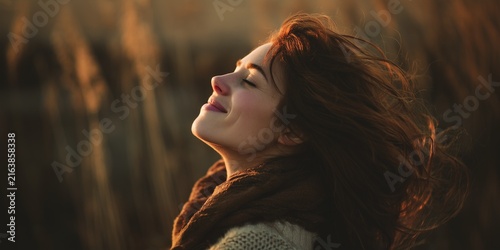 Radiant young woman, with long wavy hair, closed eyes in wind enjoying freedom, golden hour sunlight  in a soft focus field wearing a brown scarf and light wool sweater, portrait in nature