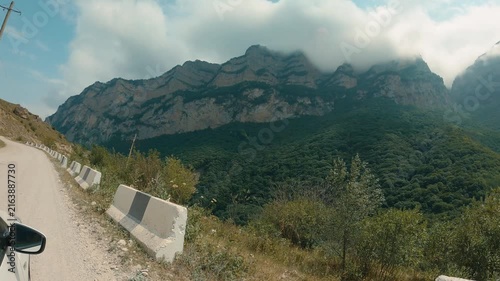 Winding dirt road curves through green valley with concrete barriers. Towering rocky mountains rise behind, partially shrouded in clouds. Blue sky with scattered white clouds
