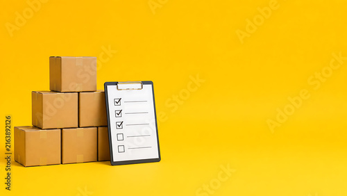 Stack of brown cardboard boxes with a clipboard showing checklist marks, symbolizing warehouse inventory management, logistics, and quality control on a solid yellow background.