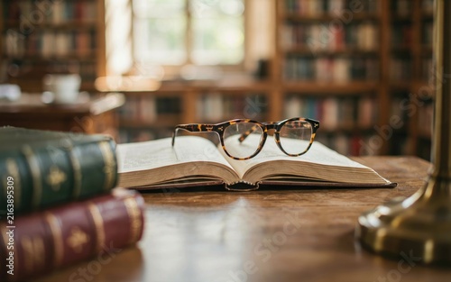 Vintage tortoise shell spectacles resting elegantly on top of an open antique hardcover book sitting on a wooden study desk