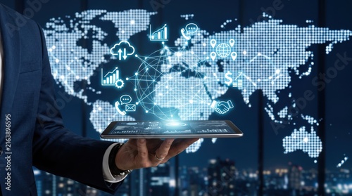 Businessman holding a tablet displaying a hologram of global network connections and digital icons over a world map, illustratings of international business, telecommunications
