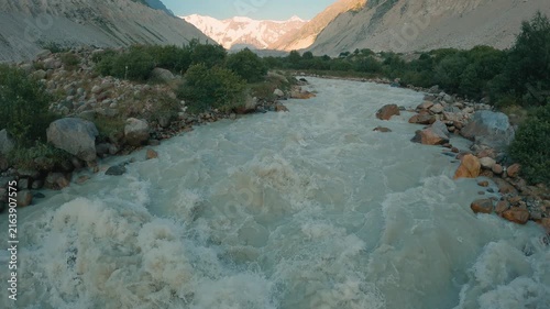 Rushing river winds through rocky valley flanked by steep slopes green shrubs. Distant snowcapped peaks under clear blue sky. Soft golden light enhances textures