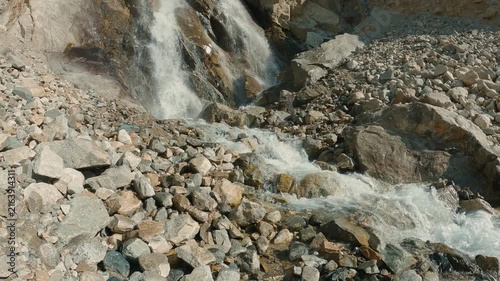 Fast-flowing water washes over rough rocks, creating a white foam. Panoramic views from the stream to the waterfall. Sunlight highlights the texture and movement