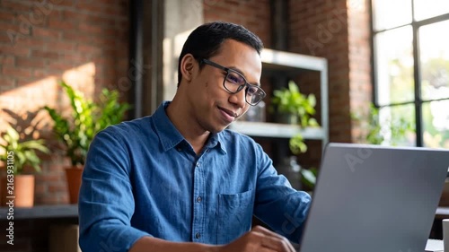 Smiling Indonesian business man looking at laptop using computer working in office. Busy professional businessman employee or company executive searching online solutions at workplace.