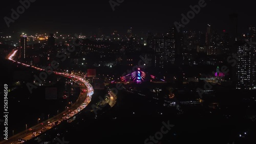 An aerial view of Mumbai's high-rise buildings and beautiful nightlights. The sky is cloudy, and there is medium traffic on the road. The cityscape is filled with modern illuminated buildings.