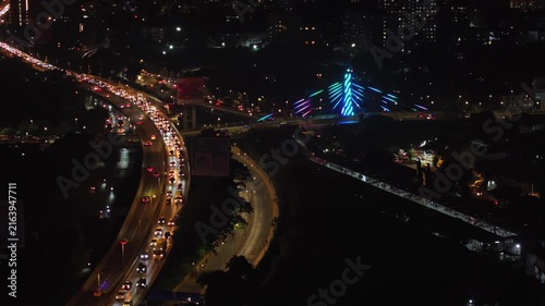 An aerial view of Mumbai's high-rise buildings and beautiful nightlights. The sky is cloudy, and there is medium traffic on the road. The cityscape is filled with modern illuminated buildings.