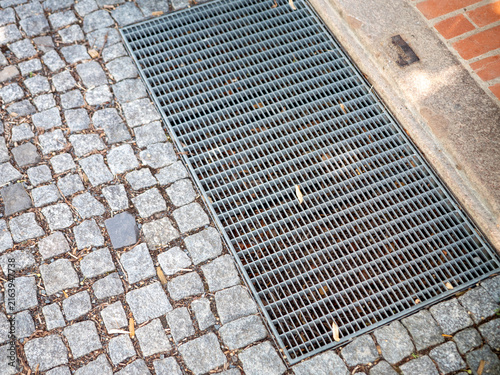 Metal drainage grate set in a cobblestone pavement at a building entrance, showing urban drainage, stone paving, grid texture and architectural detail.