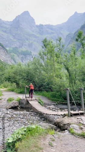 hiker crossing wooden bridge over stream in mountain valley landscape, Alps in France, vertical video